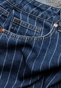 Denimstoff mit vertikalen weißen Streifen, ausgestattet mit einer Gesäßtasche, die Kupferrivets und kontrastierende Nähte aufweist.