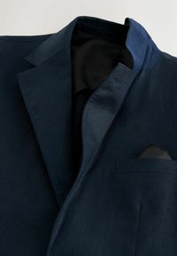 Blazer bleu marine à revers en encoche, doublure intérieure noire et pochette bleu marine assortie dans la poche poitrine.