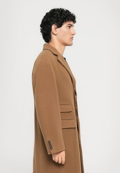 Young man with curly black hair wearing a brown overcoat, standing in profile against a plain white background.