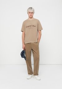 Beige cotton t-shirt with dark print, paired with beige corduroy pants. White sneakers. Model holding a dark blue cap. Simple background.
