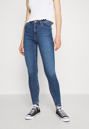 Vrouw draagt hooggetailleerde blauwe skinny jeans, witte mouwloze top met geschulpte rand en zwarte hoge sneakers, staand tegen een effen achtergrond.