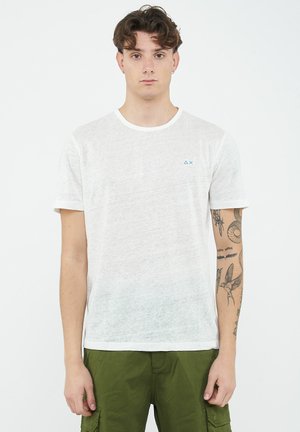 Giovane uomo con capelli castani corti che indossa una maglietta bianca e pantaloni cargo verdi, mostra i tatuaggi sull'avambraccio destro, in piedi davanti a uno sfondo bianco.