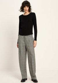 Frau mit welligem Haar, die ein schwarzes Langarmoberteil, graue karierte weite Hosen und schwarze Schuhe trägt, vor einem schlichten Hintergrund steht.