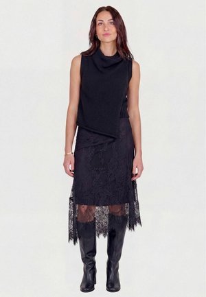 Femme portant un top asymétrique noir sans manches, une jupe en dentelle noire avec des panneaux transparents et des bottes en cuir noires montant jusqu'aux genoux sur un fond uni.