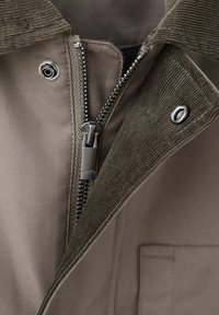 Gros plan d'une veste vert olive avec fermeture éclair métallique partiellement ouverte et deux boutons-pression sur le col en velours côtelé et la patte.