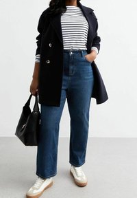 Cappotto navy doppiopetto, camicia a righe bianche, jeans in denim blu a vita alta, sneakers bianche con suola marrone e borsa nera.