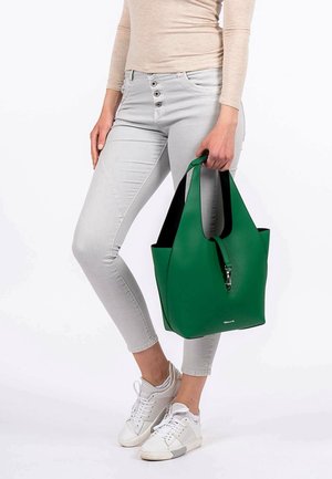 Sac fourre-tout en simili cuir vert avec un design structuré, doté d'une poignée supérieure et d'une fermeture éclair, posé sur un pantalon gris clair et des baskets blanches.