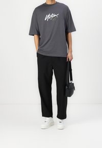 Grijze oversized t-shirt met witte grafische tekst, zwarte losse broek en witte sneakers. Een zwarte schoudertas voegt een functioneel detail toe.