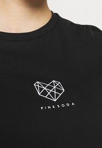 Černé tričko s bílým geometrickým logem srdce a textem "PINKSODA" pod ním. Hladká textura, kulatý výstřih, těsně přiléhající střih.