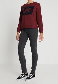 Sudadera burdeos con logo azul oscuro, combinada con jeans gris ajustados y zapatillas Vans negras, con cordones blancos y suelas de goma.