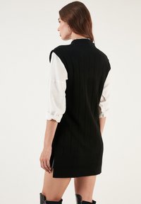 Black knitted sleeveless dress with vertical ribbed texture, worn over a white long-sleeve shirt. Features a round neckline and side slits.