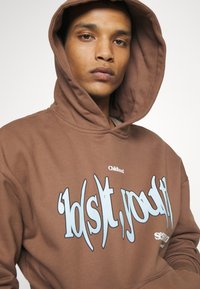 Brun hoodie med en stor blå grafisk text. Tillverkad av mjukt tyg, med dragsko i huvan och ribbade ärmslut. "Childhood"-logotypen ovanför grafiken.