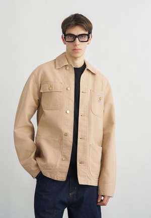 Νέος άνδρας που φοράει μαύρα γυαλιά, μπεζ σακάκι με κουμπιά και τσέπες, και σκουρόχρωμα μπλε τζιν, στέκεται μπροστά σε ένα απλό ανοιχτό φόντο.