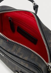 Melna rakstaina rokassoma ar sarkanu auduma oderi. Iekļauj rāvējslēdzēja aizdari un mazu iekšējo kabatu ar zīmola etiķeti. Gluda tekstūra.