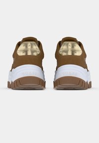 Paire de baskets marron avec semelles blanches, accents métalliques dorés au talon et marquage "GUESS" à l'arrière des semelles.