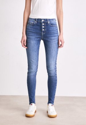 Persoon draagt een blauwe high-waist skinny jeans met vier zichtbare knopen en witte sneakers, staand tegen een effen lichte achtergrond.