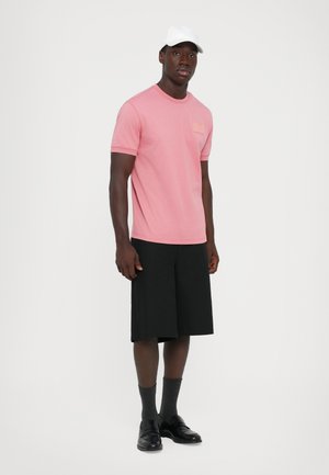 Man wearing white cap, pink short-sleeve shirt, black knee-length shorts, dark socks, and black dress shoes, standing against white background.
