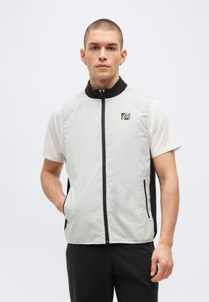 Jeune homme aux cheveux courts portant un gilet zippé sans manches gris clair par-dessus une chemise blanche à manches courtes, debout devant un fond uni.