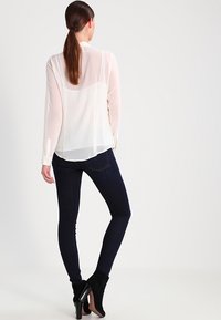 Žena s dlouhými hnědými vlasy v culíku, oblečená v průhledné bílé blůze, tmavých úzkých džínách a černých lodičkách s kotníkovým podpatekm, stojící zády.