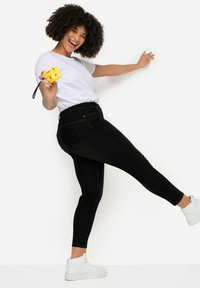 Jeans negros ajustados con bolsillos traseros, combinados con una camiseta blanca y zapatillas blancas. El sujeto sostiene una cámara amarilla, sonriendo.