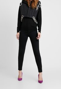 Svart kort puffjacka med huva, utrustad med vita dragskor och dekorativa etikettaccenter, ihopparad med svarta jeans och lila klackskor.