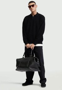 Borsa da viaggio in pelle nera con doppi manici, texture liscia e cuciture minime. Il modello indossa un polo in maglia nera e pantaloni scuri.