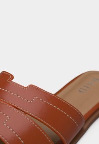 Κοντινή λήψη δερμάτινου σαγιοναρίou ταμπά με λευκές ραφές και σχέδιο με ανοίγματα στο λουράκι, σε μπεζ πάτο με το όνομα της μάρκας.