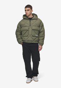 Giubbotto trapuntato verde oliva con cappuccio, dotato di due grandi tasche frontali, chiusura a mezza zip e polsini elasticizzati. Abbinato a pantaloni cargo neri.