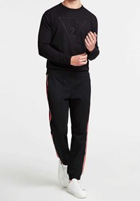 Musta huppari, jossa on kohotettu logodesign kolmiomaisessa muodossa. Yhdistetty mustiin jogger-housuihin, joissa on sivuaitoja punaisena, ja valkoisiin lenkkitossuihin.