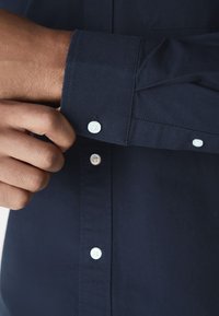 Marinblå skjorta med knappar och texturerat tyg. Har vita knappar på manschetten och knappslån samt en standardkrage.