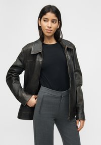 Γυναίκα με μαύρο δερμάτινο μπουφάν, μαύρο τοπ και γκρι παντελόνι, στεκόμενη με το ένα χέρι στην τσέπη, κοιτάζοντας προς τα αριστερά της, σε απλό φόντο.