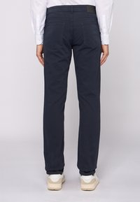 Pantalones azul marino de corte recto, confeccionados en algodón, con bolsillos traseros y una cintura plana con una etiqueta. Llevados con zapatillas blancas.