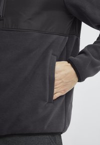 Veste en polaire gris foncé avec un panneau supérieur noir contrastant, poche latérale et poignets élastiques. Texture douce et coupe décontractée.