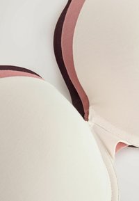 Pile de trois soutiens-gorge rembourrés en beige, rose et marron foncé, montrant un tissu lisse et des bords arrondis.