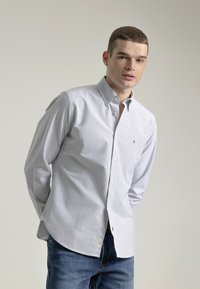 Camisa de botones a rayas en algodón azul claro y blanco. Presenta un cuello, mangas largas y una etiqueta en el dobladillo. Combinada con jeans azules.