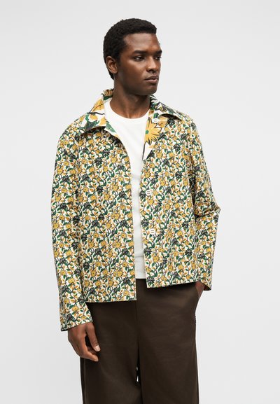 Άνδρας που φοράει κουμπωτό λουλουδένιο σακάκι με κίτρινα και πράσινα λουλούδια πάνω από λευκό πουκάμισο και καφέ παντελόνι, κοιτάζοντας δεξιά σε απλό φόντο.