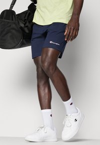 Pantalones cortos azul marino de tela con el logo de Champion, combinados con zapatillas blancas y calcetas con logo; llevando una bolsa de deporte negra.