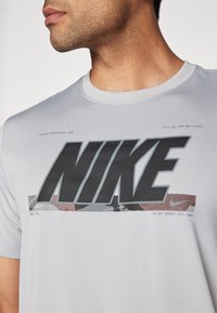 T-shirt Nike à manches courtes gris clair avec un grand logo noir et un accent en motif camouflage. Tissu doux et lisse avec un col rond.