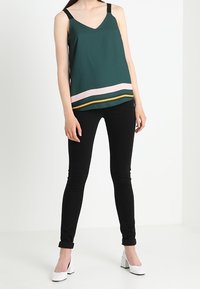 Tmavě zelený top bez rukávů s výstřihem do V a kontrastním pruhovaným okrajem v růžové a žluté barvě, kombinovaný s černými skinny džíny a bílými blokovými podpatky.