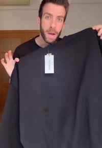 Άνδρας κρατάει μια μαύρη φούτερ με λευκή ετικέτα σε ένα δωμάτιο με ξύλινες πόρτες και τοίχους ανοιχτού χρώματος.