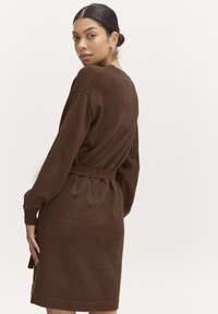 Abito in maglia marrone con maniche lunghe, vita cinte e spacchi laterali. Texture morbida, design minimale e lunghezza medio-corta.