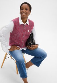 Red and blue knitted vest with buttons over a white shirt, worn with blue cropped jeans and holding a black bag, seated on a white chair.