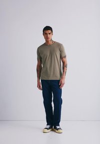 Lys olivengrønn bomulls t-skjorte med korte ermer, rund hals, sammen med mørkeblå denimjeans og svarte joggesko med kremfargede såler.