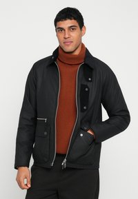 Μαύρο μπουφάν με φερμουάρ μπροστά και κουμπιά κούμπωμα, με γιακά από κορδόνι και δύο μπροστινές τσέπες; φορεμένο πάνω από ένα καφέ ριγωτό ζιβάγκο.