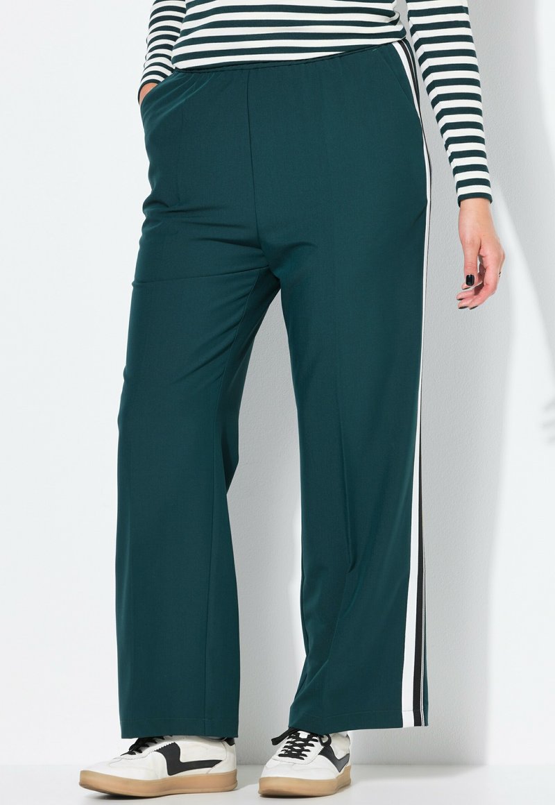 Persoon met donkergroene wijde broek met witte en zwarte zijstrepen, witte sneakers en een zwart-wit gestreept langemouwen top.