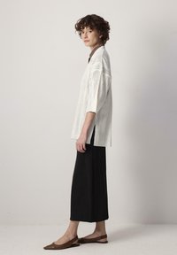 Haut tricoté blanc avec une coupe ample et des fentes sur les côtés, associé à une jupe longue noire et des ballerines pointues marron.