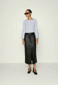 Ženska, oblečena v črne sončne očala, svetlo sivo dolgo majico z dolgimi rokavi, črno usnjeno midi krilo s sprednjim razporkom in črne masivne pete, stoji na enotni podlagi.