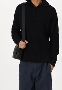 Suéter negro de tejido acanalado con cuello simulado con cremallera, combinado con un pequeño bolso cruzado negro. La modelo lleva pantalones azul marino.