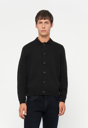 Ung mann iført en svart knappet cardigan med ribbet krage og mansjetter, kombinert med svarte jeans, stående mot en hvit bakgrunn.