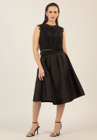 Robe noire avec un corsage en dentelle à bords festonnés, une jupe évasée et un motif brodé. Associée à des talons noirs ornés.